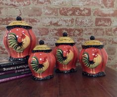 three red and yellow ceramic rooster jars on a table next to two books with brick wall in the background