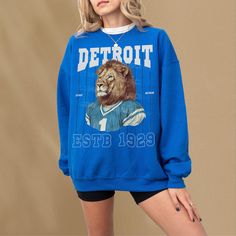 This retro Detroit football sweatshirt features a vintage illustration of a large cat wearing a blue and silver football jersey. This blue Detroit football graphic crewneck sweatshirt comes in a unsiex size for both men and women football fans. This retro football jersey sweatshirt is the perfect gift for people in Michigan who love football anybody who needs a vintage football team apparel. This sweatshirt is not associated with any team or league and is simply a piece of sports memorabilia for Retro Tops For Football Season Streetwear, Retro Tops For Streetwear During Football Season, Collegiate Graphic Print Sweatshirt For Fans, Collegiate Graphic Print Sweatshirt For Fan Merchandise, Fan Merchandise Team Colors Sweatshirt With Graphic Print, Sporty Mascot Crew Neck Tops, Blue Team Logo Sweatshirt For Sports Season, Blue Sweatshirt With Team Logo For Sports Season, Fan Merchandise Mascot Crew Neck Top