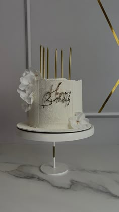 a birthday cake with gold candles and white flowers on a stand in front of a wall