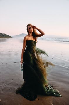 a woman in a long dress standing on the beach with her hair blowing in the wind