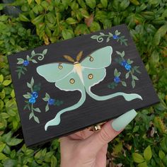 a hand holding up a painted wooden plaque with a butterfly on it's wings