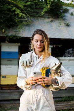 a woman in white overalls holding a cell phone and beekeeper's gloves