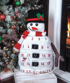 a crocheted snowman sitting next to a christmas tree in front of a fireplace