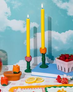 two yellow candles sitting on top of a table next to some fruit and other items