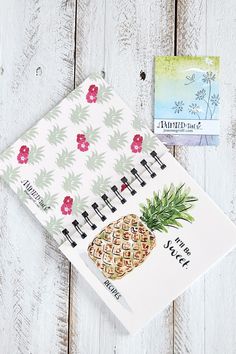 a pineapple themed notebook next to an empty notepad on a white wooden surface