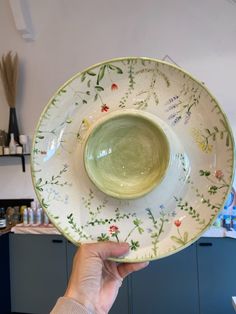 a person holding up a green and white bowl with flowers on the rim, in front of a blue cabinet