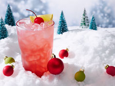 a red drink in a glass surrounded by christmas ornaments and pine cones on top of snow