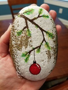 a hand holding a painted rock with a tree branch and ornament on it