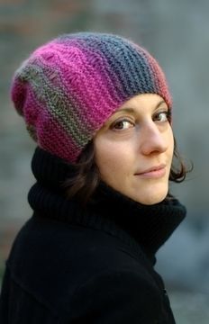 a woman wearing a knitted hat and black coat looks up at the camera while standing in front of a brick wall