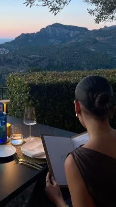 a woman sitting at an outdoor table with wine glasses and plates on it, overlooking the mountains