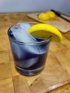 a glass filled with ice and a lemon wedge on top of a wooden table next to a cutting board