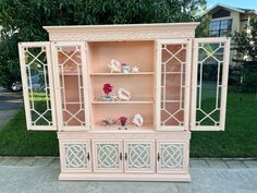 an old china cabinet painted pink with white trim and flowers on the top shelf, sitting in front of a house