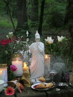 a table with candles, flowers and statues on it in the middle of a forest