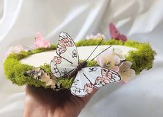 a hand holding a white butterfly on top of a green moss covered hat with pink and white flowers