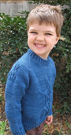 a young boy wearing a blue sweater and brown pants standing in front of some bushes