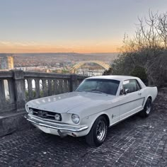 an old white mustang parked on the side of a stone road in front of a cityscape