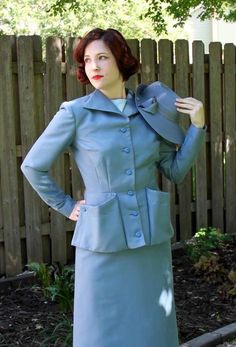 a woman in a blue dress and jacket standing next to a fence with her hands on her hips