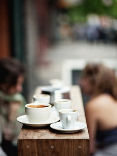 three cups of coffee sit on a wooden table with two people sitting in the background