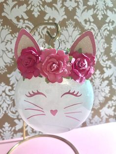 a white cat with pink flowers in it's head is on a stand next to a wall