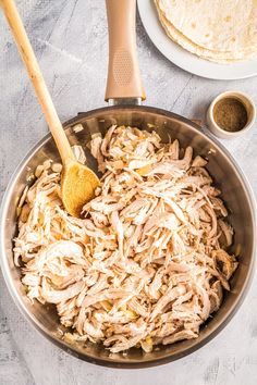 shredded chicken in a pan with a wooden spoon next to tortilla shells and seasoning