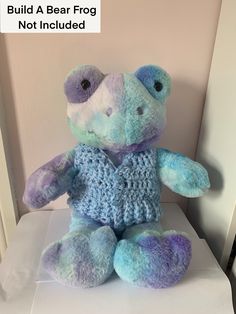 a blue and purple teddy bear sitting on top of a white box with the words build a bear frog not included