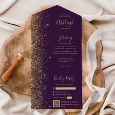 a purple and gold wedding card on top of a wooden slab with white fabric around it