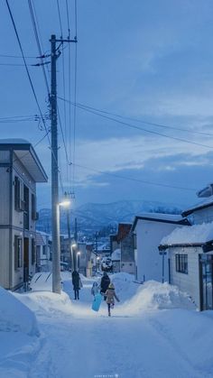 two people walking down a snow covered street in the middle of town at night time