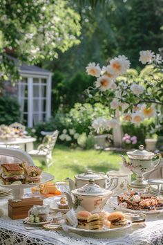 a table with tea and sandwiches on it in the middle of a garden filled with flowers