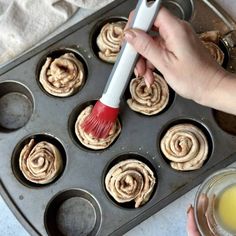 a person holding a red brush in a muffin tin filled with cinnamon buns