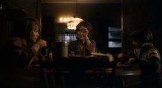 three women sitting at a table in a dark room, one reading a book and the other talking