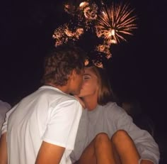 a man and woman sitting next to each other with fireworks in the sky behind them