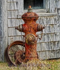 an old rusted fire hydrant sitting in front of a wooden building next to a wheel