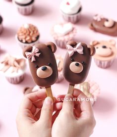 someone is holding two chocolate teddy bear lollipops in front of cupcakes