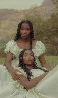 two women sitting on the ground with trees in the background, one wearing a white dress