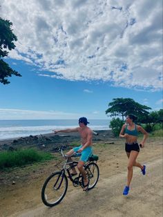 two people running and riding bikes on a dirt road next to the ocean under a cloudy blue sky