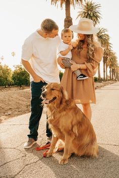 a man and woman holding a baby standing next to a brown dog on the sidewalk