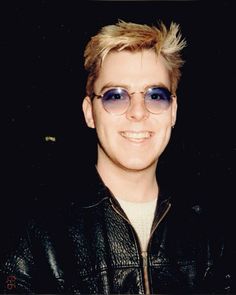 a man with blonde hair wearing sunglasses and smiling at the camera while standing in front of a black background