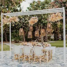 an outdoor dining set up with white chairs and flowers hanging from the ceiling, surrounded by greenery