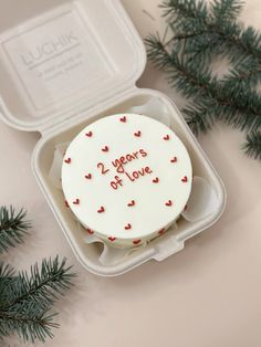 a white cake with red hearts on it in a plastic container next to pine branches