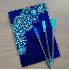 two pens sitting on top of a blue notebook next to another pen and paper with an intricate design