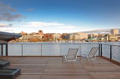 two chairs sitting on top of a wooden deck next to the ocean with city skyline in the background