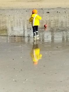 a small child is walking on the beach with a ball in his hand and wearing a yellow jacket