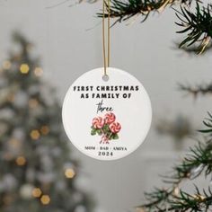 a white ornament hanging from a christmas tree with pink roses on it and the words first christmas as family of three