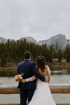 a bride and groom standing on a bridge looking at the lake in front of them