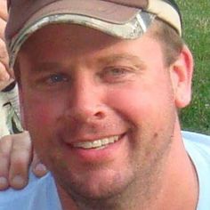 a close up of a person wearing a hat and smiling at the camera with grass in the background