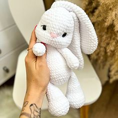 a hand holding a white crocheted bunny toy in front of a chair with a tattoo on it's arm