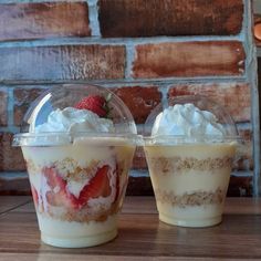 two desserts with strawberries and whipped cream in plastic cups on a wooden table