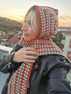 a woman wearing a crocheted hat and scarf on top of a roof with the sun setting in the background