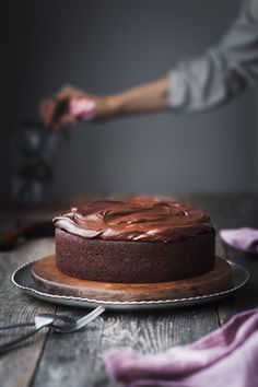 a chocolate cake with frosting being cut on a wooden table next to utensils