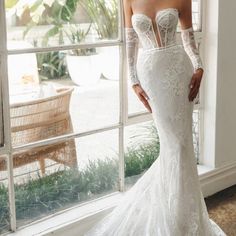 a woman in a wedding dress standing by a window with her hands on her hips